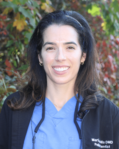 Dr. Gabriella Marti, DMD, smiling in a dental office setting wearing scrubs and a white coat at Pediatric Dentistry & Orthodontics of Salem in Salem, NH